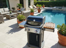 A BBQ Boards® Weber Spirit Old Gen Side Cutting Board sits on a modern black and silver gas grill near a poolside patio, surrounded by potted plants and wicker outdoor furniture with cushions in a bright, summery setting.