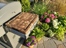A BBQ Boards® Weber Spirit Old Gen Side Cutting Board rests on a grill’s side shelf, surrounded by potted plants and blooming flowers on a sunlit stone patio.