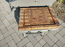 A BBQ Boards® Weber Spirit Old Gen Side Cutting Board with handles rests on a gray grill's side shelf, its wood grain and engraved logo visible in the sunlight—a practical prep station accessory for outdoor grilling.