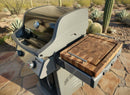 A Weber Spirit grill with a BBQ Boards® Weber Spirit Old Gen Side Cutting Board sits on a stone patio surrounded by desert plants and cacti, making outdoor meal prep easy.