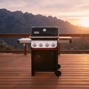 A BBQ Boards® Weber Spirit Reinvented Side Cutting Board is displayed on a black and silver Weber Spirit grill at sunset, set on a wooden deck with mountains and a scenic valley in the background.