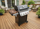 A BBQ Boards® Weber Spirit Reinvented Side Cutting Board is placed on a black gas grill with three knobs, set on a wooden deck among patio chairs and potted plants, with a house and lush greenery in the background.