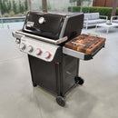 A black Weber Spirit Reinvented gas grill with four dials and a lid thermometer features the BBQ Boards® Weber Spirit Reinvented Side Cutting Board on its side board, set on a patio with outdoor seating and greenery in the background.