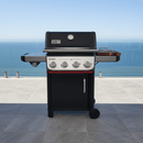 A black gas grill with four control knobs and side shelves stands on a tiled patio near the ocean, featuring the BBQ Boards® Weber Spirit Reinvented Side Cutting Board by BBQ Boards® for convenient prep.