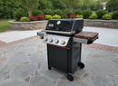 A BBQ Boards® Weber Spirit Reinvented Side Cutting Board is shown attached to a black Weber Spirit grill, featuring three control knobs and side shelves, set on a stone patio in a landscaped yard with bushes and flowers.