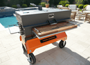 A BBQ Boards® Yoder Flat Top Front Cutting Board by BBQ Boards® is set on a large black and orange Yoder smoker grill with a griddle and wooden shelf, positioned on a patio near wicker outdoor furniture and lush greenery beside a pool.