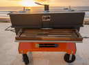 A BBQ Boards® Yoder Flat Top Front Cutting Board is attached to a barbecue smoker on a tiled patio by the ocean at sunset, with sunlight reflecting off the water in the background.