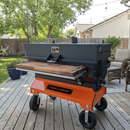 The BBQ Boards® Yoder Flat Top Front Cutting Board by BBQ Boards® is shown on a modern smoker grill with an orange base and wood shelf, set on a backyard deck with patio furniture, greenery, and string lights above.