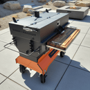 A large black and orange outdoor Yoder smoker with a wooden side shelf features the BBQ Boards® Yoder Flat Top Front Cutting Board, providing generous griddle space on a patio with rocks and a modern fire pit in the background.