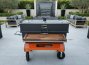 BBQ Boards® Yoder Flat Top Front Cutting Board on a Yoder Smokers charcoal smoker with orange base, wood shelf, and gray cooking chamber on a patio, surrounded by gray outdoor sofas, stone water feature, and lush greenery.