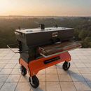 The BBQ Boards® Yoder Flat Top Front Cutting Board by BBQ Boards® is on a large outdoor smoker grill with a griddle on wheels, set on a tile patio overlooking scenic trees and hills at sunrise or sunset.