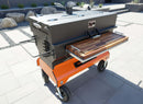 A large, rectangular outdoor smoker grill on wheels features a black top, orange base, BBQ Boards® Yoder Flat Top Front Cutting Board for extra griddle workspace, wooden side shelf, and metal handles; set on a patio with rocks and plants in the background.