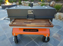 A large outdoor barbecue smoker with a black top, BBQ Boards® Yoder Flat Top Front Cutting Board, wooden front shelf, and orange wheeled base sits on a stone patio with a fire pit and landscaping in the background.