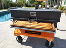 A large Yoder Smokers grill with an orange base and a BBQ Boards® Yoder Flat Top Front Cutting Board stands on a patio by a pool, amid palm trees and outdoor furniture, offering the ideal griddle workspace for outdoor cooking.