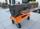A Yoder Smokers charcoal grill with a BBQ Boards® Yoder Flat Top Front Cutting Board and built-in wooden side shelf sits on a modern patio by the pool. The grill has large wheels, an orange base, and outdoor seating visible in the background.