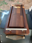 A pair of BBQ Boards® Yoder YS1500 Front & Pellet Bin Cutting Boards by BBQ Boards® with metal handles rest atop a grill above a coil handle, set against dirt and shaded outdoors—perfect for BBQ prep.