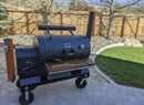 A large black outdoor smoker grill sits on a stone patio, featuring a handy BBQ prep station with BBQ Boards® Yoder YS1500 Front & Pellet Bin Cutting Boards (Pair) by BBQ Boards®, plus space for accessories in the fenced backyard.