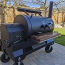 BBQ Boards® Yoder YS1500 Front & Pellet Bin Cutting Boards (Pair) from BBQ Boards® are shown on a large black outdoor smoker grill with a chimney, metal handles, wooden shelf, and wheels on a stone patio near a wooden fence.
