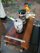The BBQ Boards® Yoder YS1500 Front & Pellet Bin Cutting Boards (Pair) from BBQ Boards® serve as a convenient BBQ prep station, holding a beer and barbecue sauces on an outdoor grill with greenery in the background.