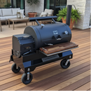 A large black outdoor smoker grill with wheels is on a wooden deck near patio furniture, potted plants, and a BBQ prep station featuring BBQ Boards® Yoder YS1500 Front & Pellet Bin Cutting Boards (Pair) by BBQ Boards®, in front of a modern house.