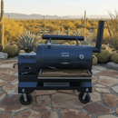 A BBQ Boards® Yoder YS1500 Front Cutting Board is set on a black smoker grill atop a stone patio in a desert scene, with cacti and mountains in the background under a clear sky.