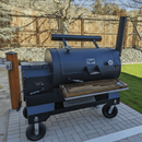 The BBQ Boards® Yoder YS1500 Front Cutting Board by BBQ Boards® adds ample prep space to a large black smoker grill with chimney and temperature gauge, set on a patio with a wooden fence and trees in the background.