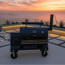 A BBQ Boards® Yoder YS1500 Front Cutting Board by BBQ Boards® provides convenient prep space on a black outdoor smoker grill overlooking the ocean at sunset, with geometric tiles, modern planters, and warm lighting in the background.