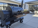 A BBQ Boards® Yoder YS1500 Front Cutting Board by BBQ Boards® is set on a large barrel smoker grill with a wooden side shelf on a modern patio, backed by outdoor seating and a contemporary house with big windows and landscaping.