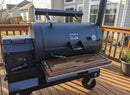 A BBQ Boards® Yoder YS1500 Front Cutting Board by BBQ Boards® is featured on a large black outdoor smoker grill with a built-in thermometer and spacious wooden prep shelf, set on a deck with houses and plants in the background.