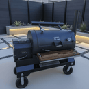 A black outdoor smoker grill with a BBQ Boards® Yoder YS1500 Front Cutting Board stands on wheels on a modern patio with concrete tiles, gravel, desert plants in raised beds, and a dark privacy fence.