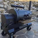 A large black Yoder smoker grill with a side firebox and wooden shelf stands on a stone patio, featuring ample prep space and a sturdy BBQ Boards® Yoder YS1500 Front Cutting Board, with terraced rock landscaping in the background.