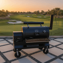 A black barbecue smoker with a BBQ Boards® Yoder YS1500 Front Cutting Board on wheels stands on a tiled patio, overlooking a manicured golf course and trees beneath a warm sunset sky.