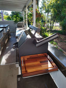 A black grill with a closed lid sits on a patio countertop, featuring the BBQ Boards® Yoder YS480 or YS640 Side Cutting Board as the perfect BBQ prep surface. Sunlight casts shadows over lush greenery while a wooden cutting board rests on the shelf.