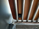 Close-up of a metal and wood structure, featuring vertical metal rods, a BBQ Boards® Yoder YS480 or YS640 Side Cutting Board, and a black screw securing them. The background is out of focus.