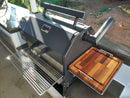 A black outdoor smoker grill features the BBQ Boards® Yoder YS480 or YS640 Side Cutting Board on its side shelf, set on a patio with sunlight and greenery in the background.