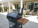 A BBQ Boards® Yoder YS480 or YS640 Side Cutting Board is on a black outdoor smoker grill set up on a sunny backyard patio with covered seating, tables, chairs, and potted plants.