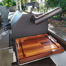A black outdoor smoker grill with a spacious BBQ prep surface features a BBQ Boards® Yoder YS480 or YS640 Side Cutting Board attached. Sunlight brightens the area, which is surrounded by lush greenery.