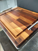 The BBQ Boards® Yoder YS480 or YS640 Side Cutting Board, featuring handles and a striped wood pattern, sits on the counter—ideal as a BBQ prep surface or as a stylish side board for your grill.