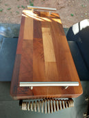 A wooden grill press rests on BBQ Boards® Yoder YS480 Trio Front, Side & Pellet Bin Cutting Boards Set of Three, with sunlight casting shadows across the BBQ Boards® surfaces and textured press.