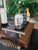 A close-up of the BBQ Boards® Yoder YS480 Trio Front, Side & Pellet Bin Cutting Boards Set of Three holding a can of Stone beer, BBQ rub, and sauce on an outdoor grill prep station amid green foliage.