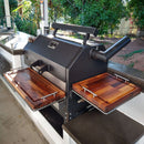 A Yoder YS640 smoker grill is outfitted with BBQ Boards® Trio Front, Side & Pellet Bin Cutting Boards (Set of 3) by BBQ Boards®, positioned on a countertop in a covered patio with trees and greenery in the background.