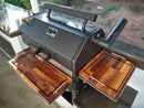 A black Yoder YS640 grill with BBQ Boards® Yoder YS640 Trio Front, Side & Pellet Bin Cutting Boards (Set of 3) sits on a patio countertop, providing expanded workspace with a yard and greenery in the background.