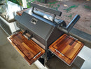 A Yoder YS640 grill is outfitted with BBQ Boards® Trio Front, Side & Pellet Bin Cutting Boards (Set of 3) on a patio, surrounded by stainless steel countertops and a lush garden—delivering the ultimate grill workspace upgrade.