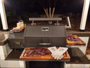 At night, smoked meat rests on BBQ Boards® Yoder YS640 Trio Cutting Boards (Set of 3) by BBQ Boards®, with barbecue gloves and seasonings nearby, upgrading a grill workspace in an outdoor kitchen. Trees and a fence are seen in the background.