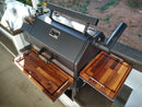 The BBQ Boards® Yoder YS640 Trio Front, Side & Pellet Bin Cutting Boards (Set of 3) by BBQ Boards® are shown on wooden side shelves of a black outdoor smoker, enhancing your grill workspace on a patio with kitchen supplies nearby.