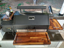 A large black Yoder YS640 grill features BBQ Boards® Yoder YS640 Trio Front, Side & Pellet Bin Cutting Boards (Set of 3) as shelves, holding a can, sauce bottle, and beer bottle on the left shelf, set on a patio with greenery behind.