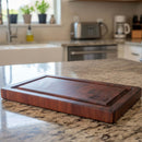 The BBQ Boards® Basic BBQ Board® 16" x 9" Cutting Board rests on a granite kitchen countertop, with a sink, potted plants, toaster, and coffee maker blurred in the background.