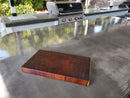 The BBQ Boards® Basic BBQ Board® 16" x 9" cutting board, made of dark wood, rests on a shiny outdoor kitchen countertop with a grill and sink behind it and green foliage visible beyond the counter.