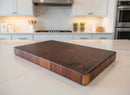 The Basic BBQ Board® 16" x 9" Cutting Board by BBQ Boards® features an engraved BBQ-themed logo on one corner, shown on a white marble kitchen island with a modern kitchen backdrop.