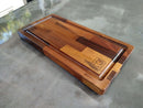 The Beast BBQ Board® 20" x 10" Cutting Board by BBQ Boards® features a patchwork wood design, edge groove, and “BBQ Chops” with a pig graphic engraved in one corner. It sits on a gray countertop.
